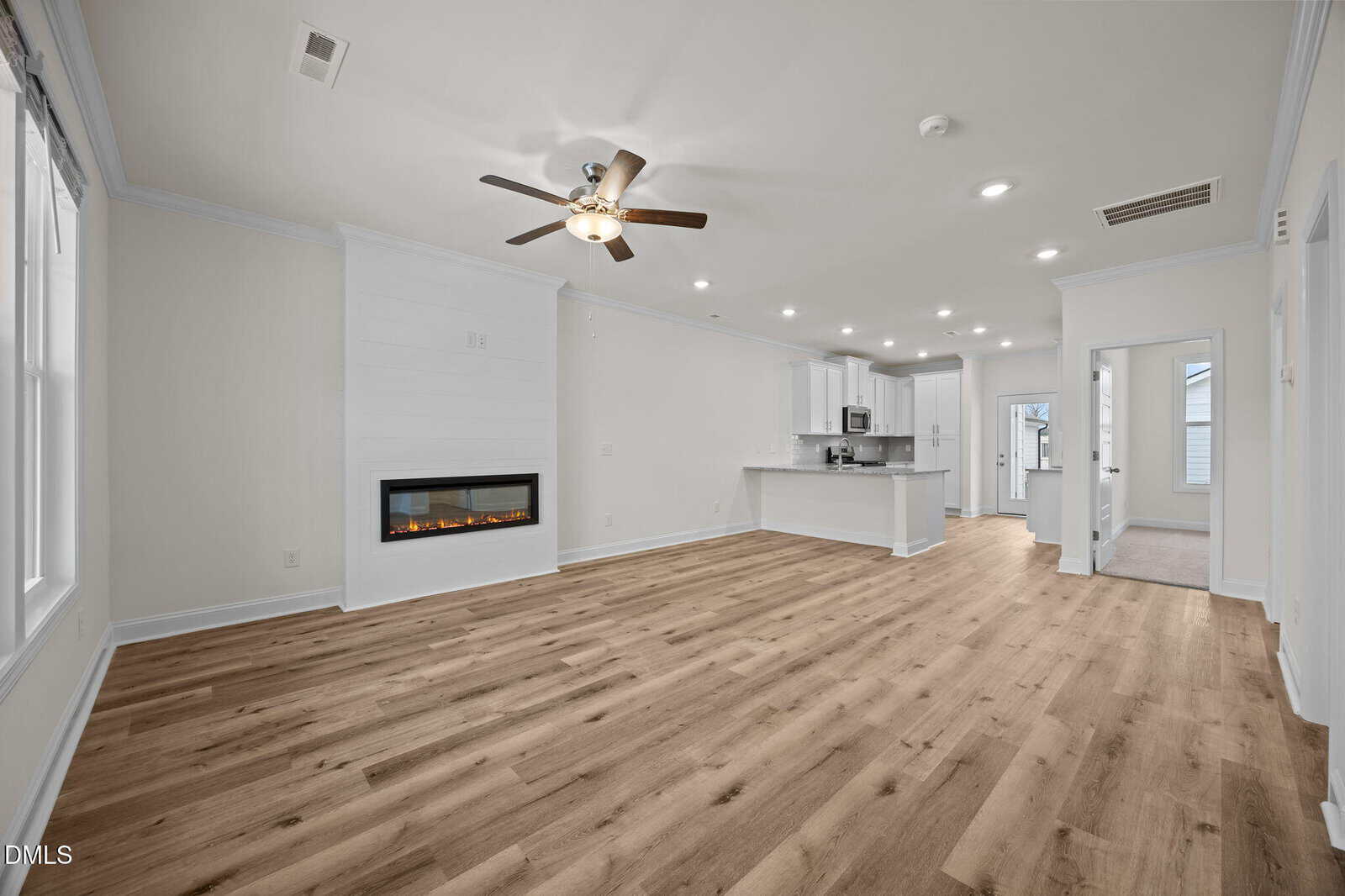 Open-concept living space with wood flooring, white walls, a ceiling fan, wall-mounted fireplace, and a modern kitchen with white cabinets and an island in the background. Large windows provide natural light.