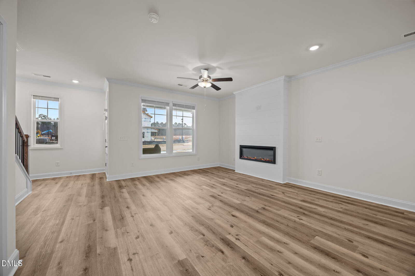 Spacious, modern living room with light wood flooring, white walls, a ceiling fan with lights, recessed lighting, a wall-mounted electric fireplace, and large windows letting in natural light.