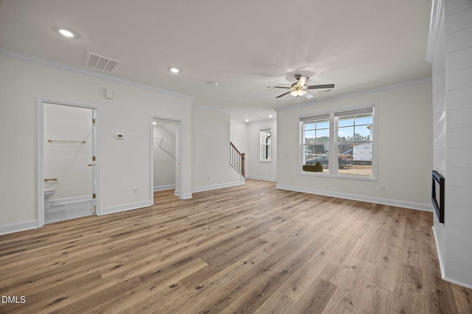 Spacious living room with light wood flooring, white walls, ceiling fan, large front windows, entry to a bathroom on the left, stairs in the back, and sunlight coming through the windows.
