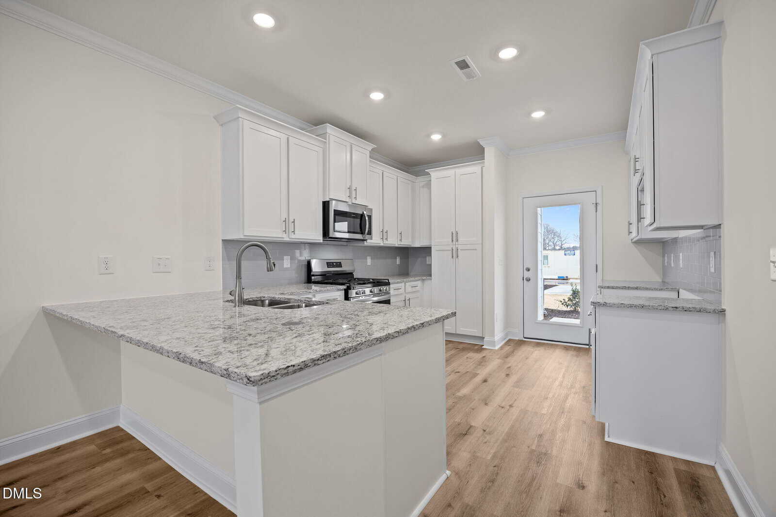 Modern kitchen with white cabinets, stainless steel appliances, granite countertops, a center island with a sink, and light wood flooring. Recessed lighting brightens the space, and a glass door leads outside.