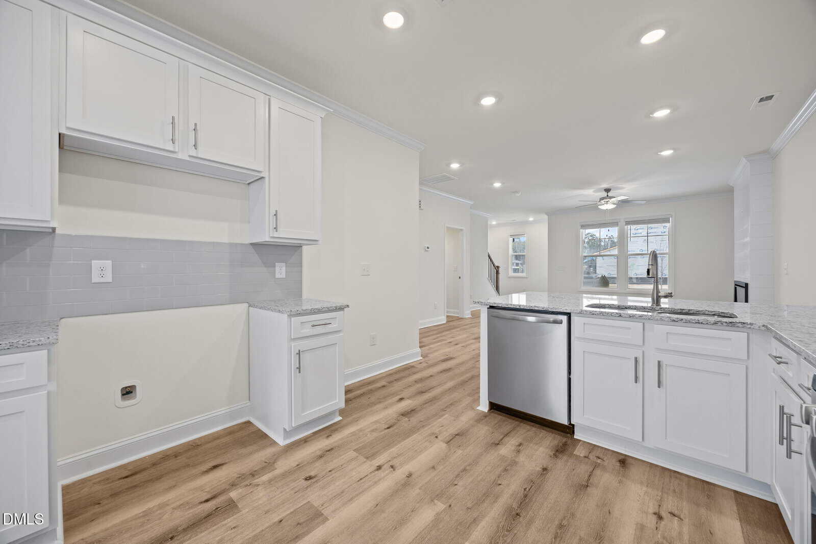 Modern kitchen with white cabinets, granite countertops, stainless steel appliances, light wood flooring, and open layout leading to a bright living area with large windows and ceiling fan.