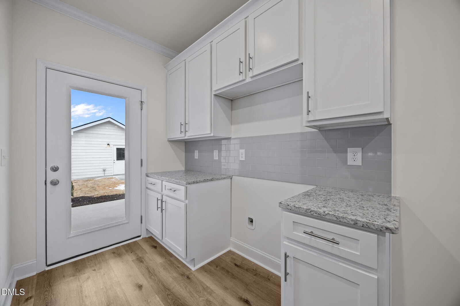 A bright laundry or utility room with white cabinets, granite countertops, light gray tile backsplash, and light wood flooring. A door with a window opens to an outdoor area with a white building.