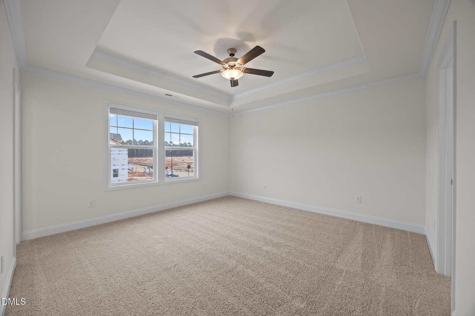 A spacious, empty bedroom with beige carpet, white walls, a tray ceiling with a ceiling fan, two large windows letting in natural light, and a view of a construction area outside.