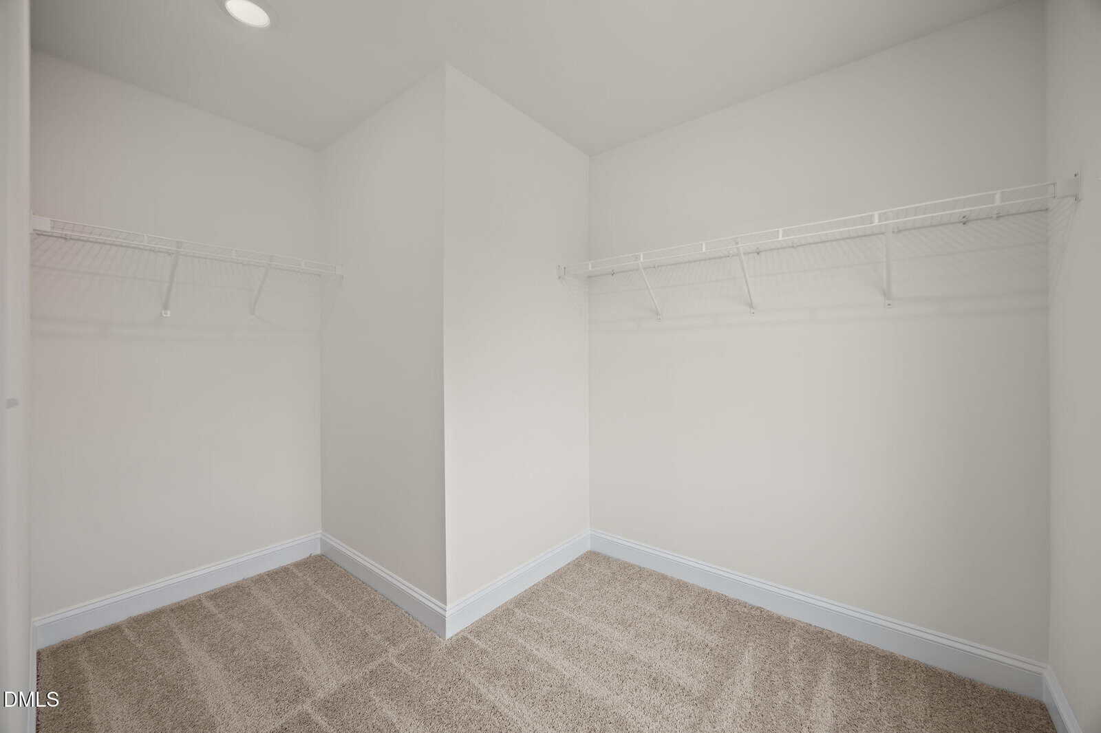 Empty walk-in closet with beige carpet, white walls, and two white wire shelves on either side. The space is brightly lit and has a clean, modern appearance.