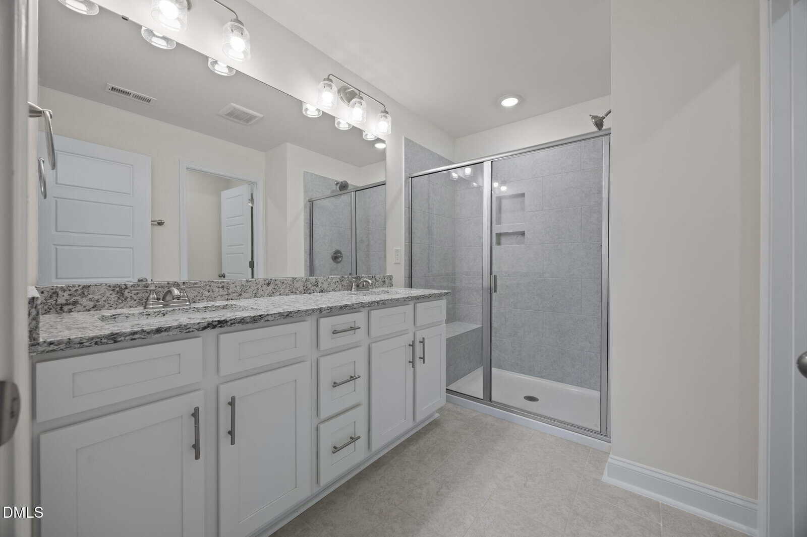 Modern bathroom with double sink vanity, granite countertop, large mirror, and a glass-enclosed walk-in shower. Walls and cabinets are white, and the floor is light-colored tile. Bright lighting above the mirror.