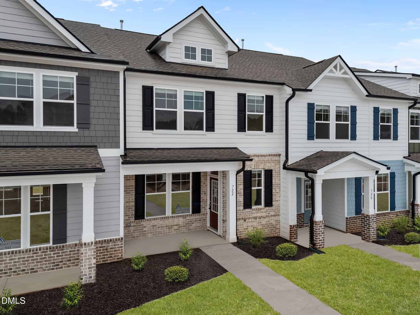A modern two-story townhouse with gray and white siding, brick accents, large windows, a covered front entry, and neatly landscaped shrubs and grass.