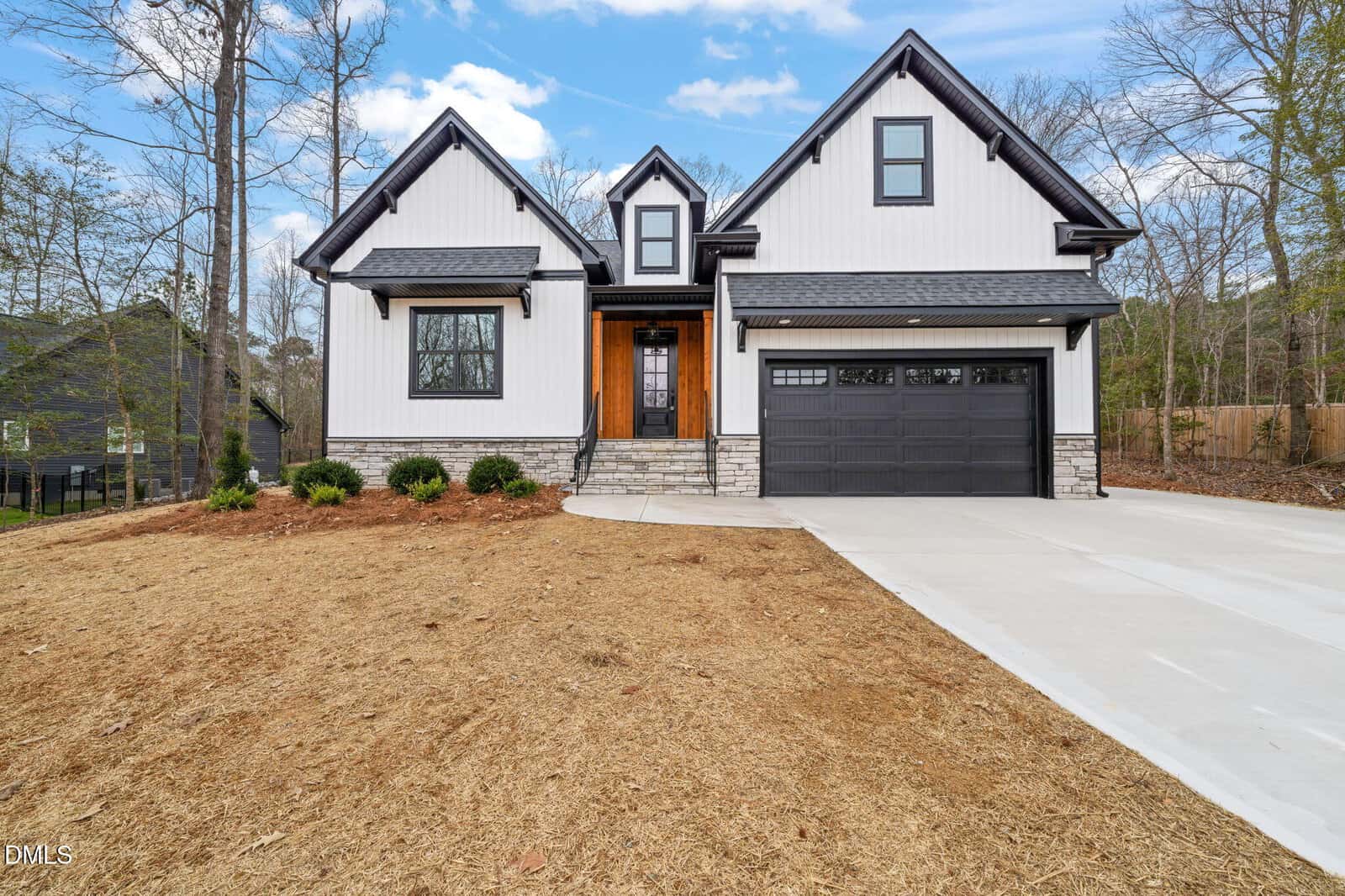 Modern two-story house with white board-and-batten siding, black trim, a double garage, stone accents, and a concrete driveway. The yard is mostly bare with a few shrubs near the entry. Trees and neighboring homes are visible.
