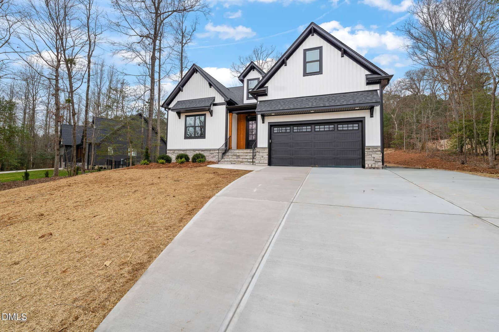 Modern two-story house with white siding, black trim, and a dark gray double garage door, situated on a large sloped lot with a wide concrete driveway and surrounded by leafless trees.