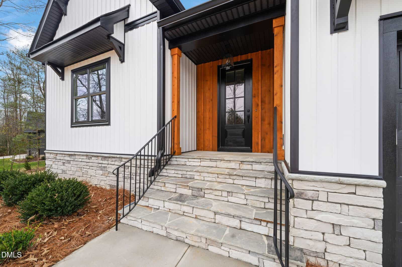 A modern house entrance with stone steps, black railing, and a black front door framed by natural wood columns, white vertical siding, and black-trimmed windows. Shrubs and mulch line the walkway.