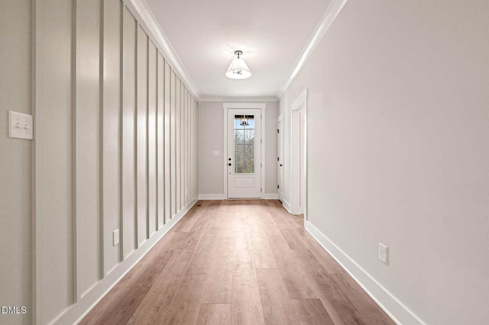 Bright hallway with light wood flooring, white trim, paneled wall on the left, and a glass door at the far end letting in natural light. The walls are painted a soft, neutral color.