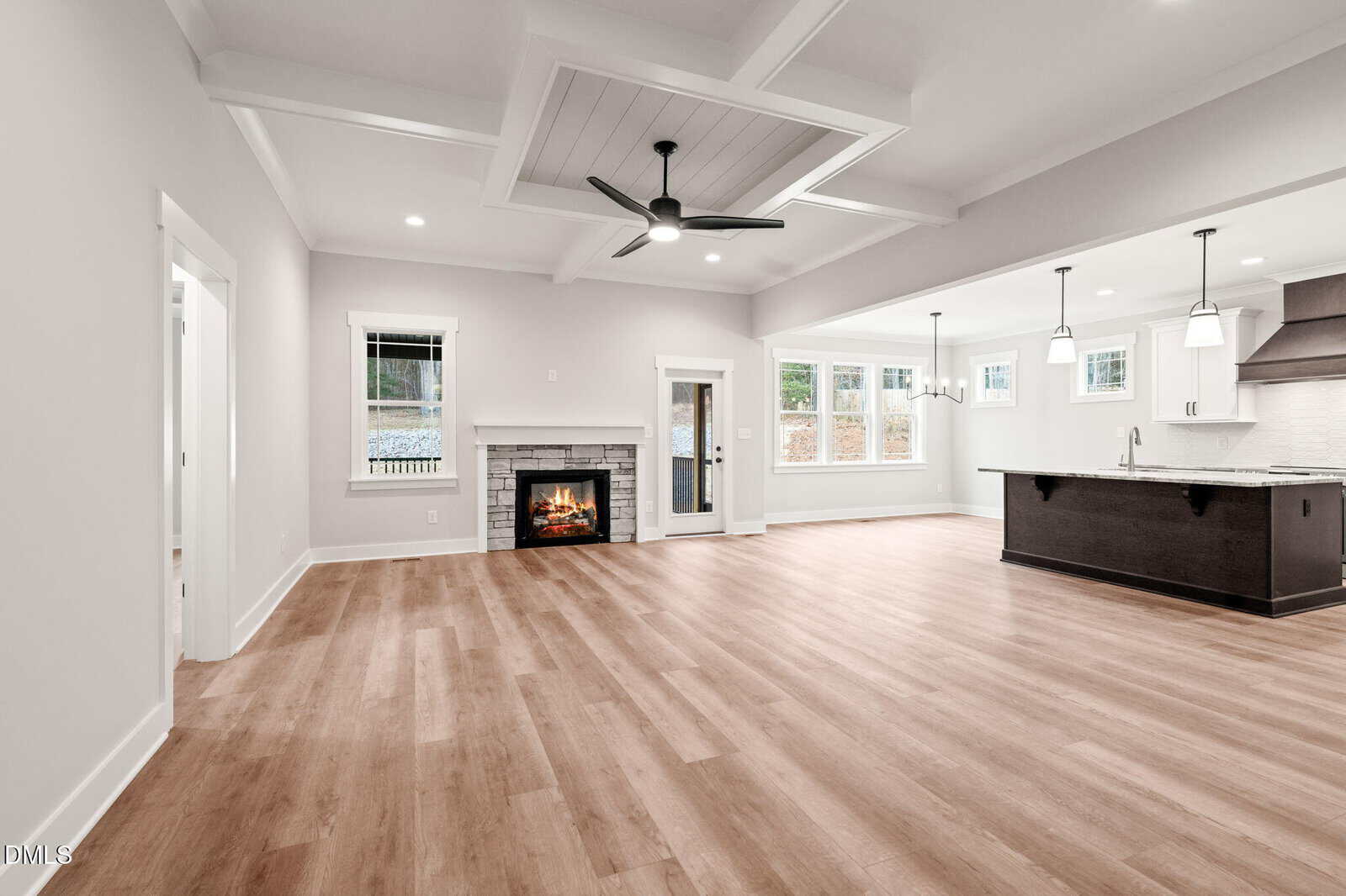 Spacious, modern open-concept living area with light wood flooring, a ceiling fan, fireplace, and large windows. The kitchen features a dark island, white cabinetry, pendant lights, and stainless steel appliances.