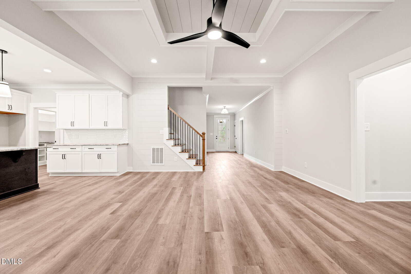 Spacious modern interior with light wood flooring, white walls, recessed lighting, ceiling fan, open kitchen with white cabinets, and a staircase leading upstairs. Hallway extends toward the front door.
