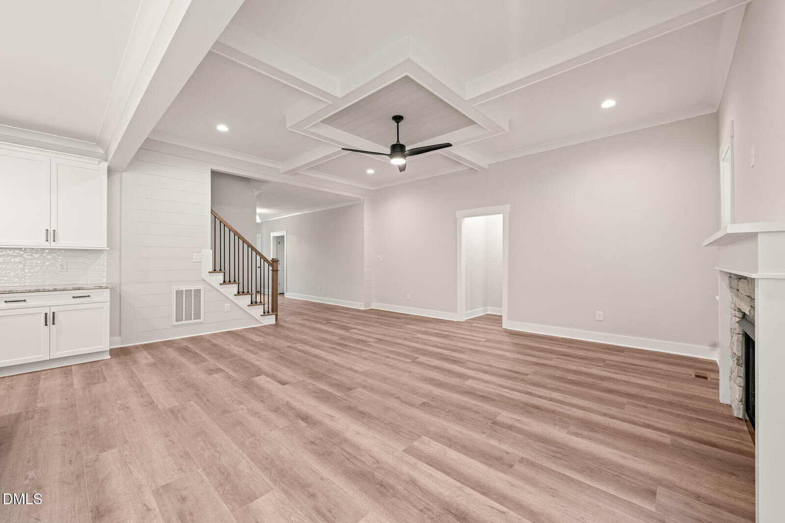 Spacious, modern living room with light wood floors, white walls, ceiling fan, coffered ceiling, and a fireplace. Open layout connects to a white kitchen and a staircase with wooden handrails.