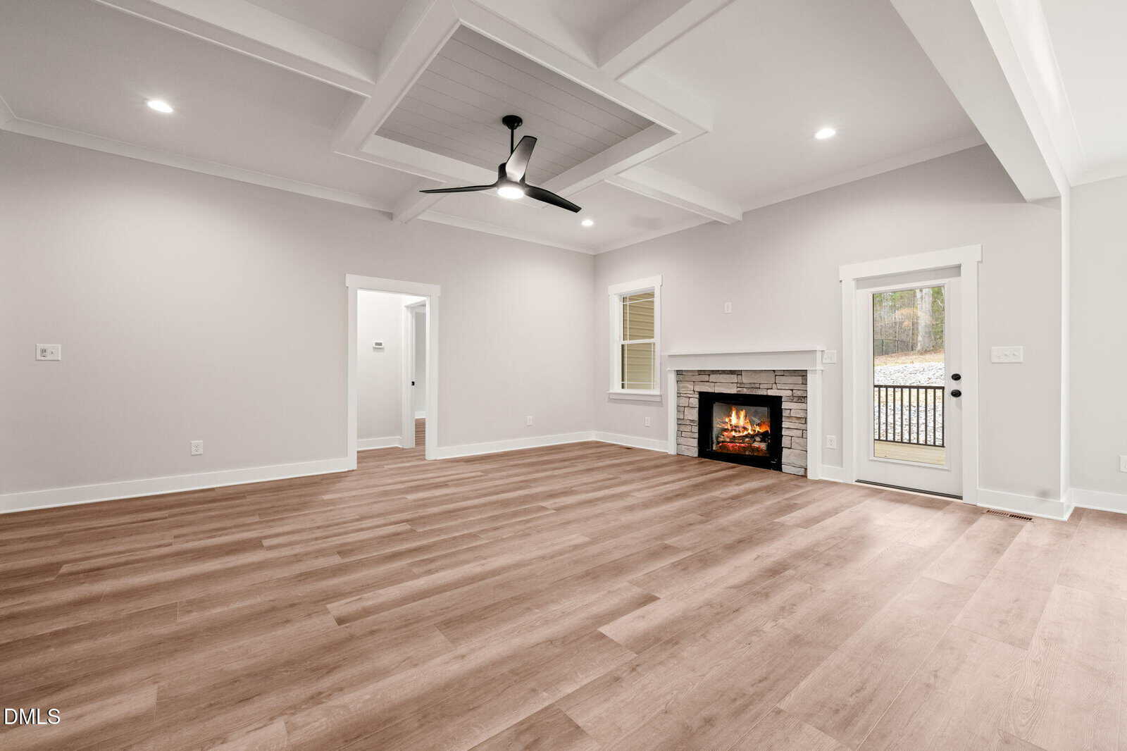 Spacious modern living room with light wood flooring, a coffered ceiling with a black fan, a fireplace with a stone surround, and white walls. There is a glass door leading outside and several doorways.