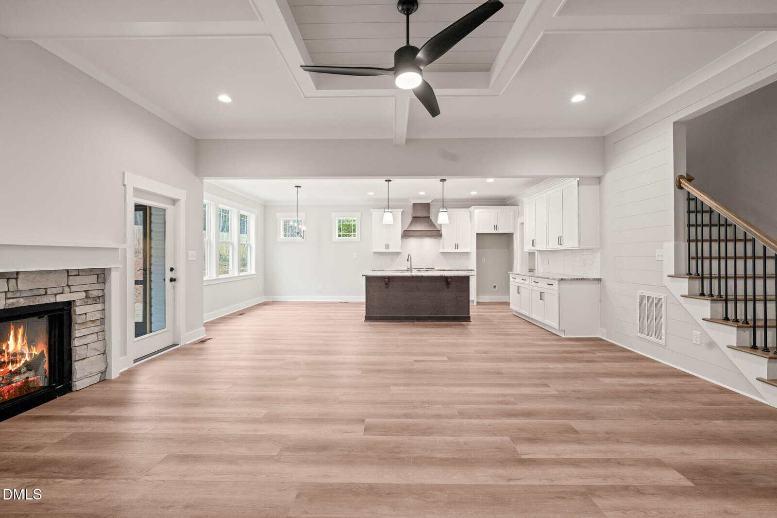 Spacious modern open-concept living area with light wood floors, a stone fireplace, large kitchen island, white cabinets, stainless steel appliances, and a ceiling fan. Stairs are visible to the right.