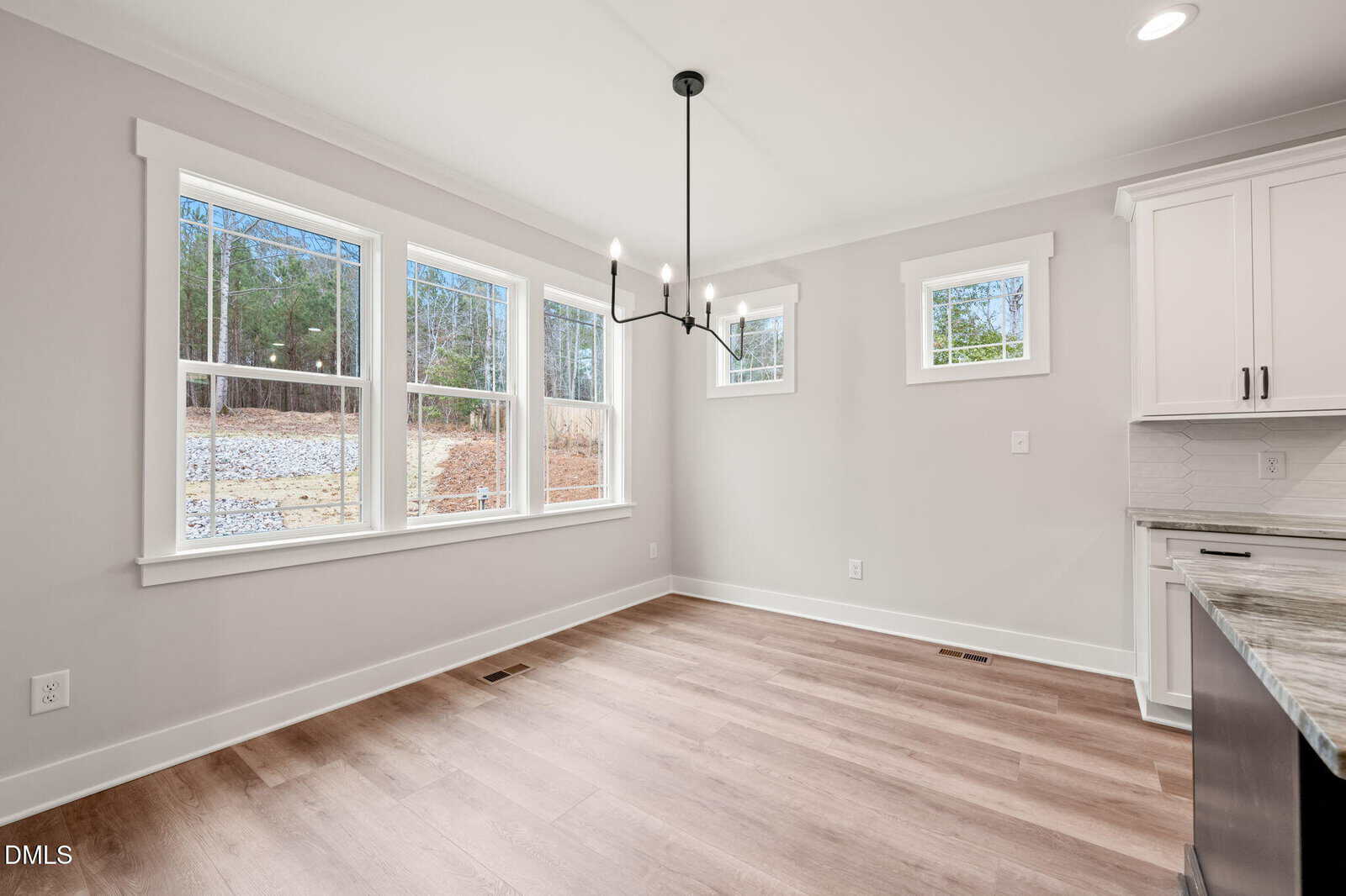 Bright dining area with large windows, light wood floors, a modern black chandelier, and white walls, adjacent to a kitchen with white cabinets and marble countertops.