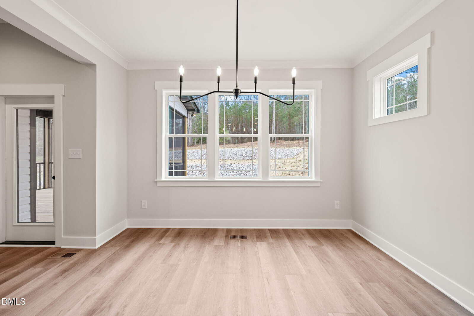 A bright, empty dining room with light wood floors, large windows facing a wooded yard, white walls, and a modern black chandelier hanging from the ceiling.