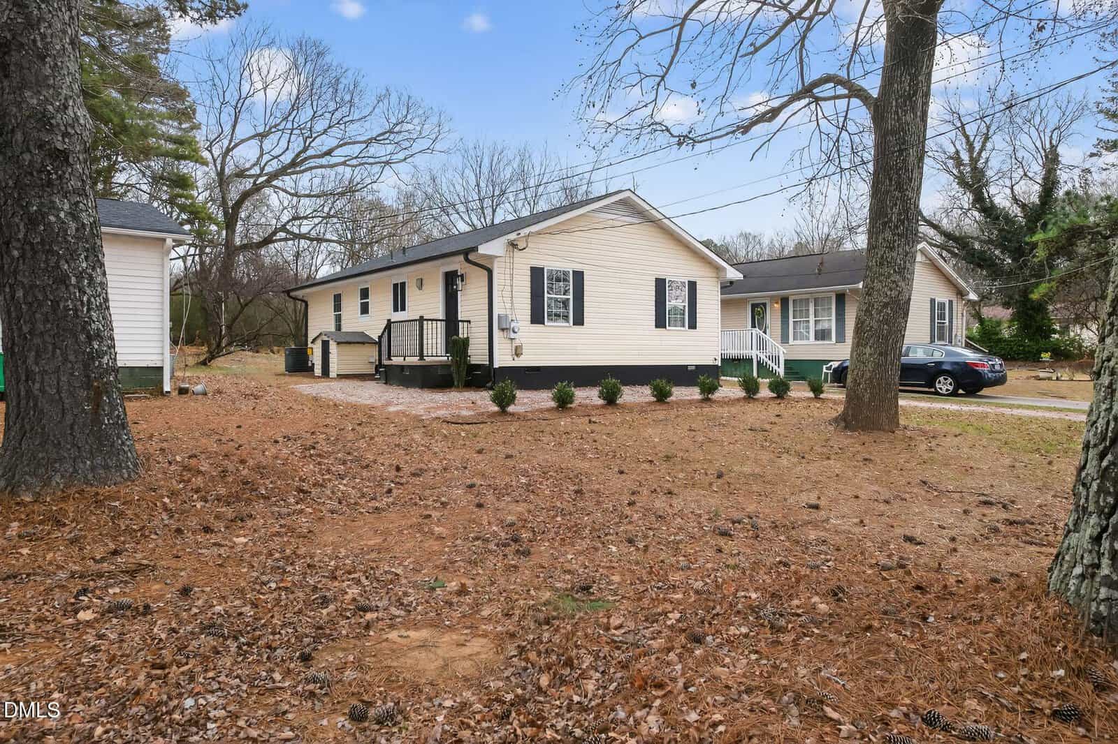 A single-story beige house at 215 Wilmington Avenue in Oxford, featuring black shutters and a small front porch, is surrounded by trees and a yard covered in pine needles. Another green-sided house sits in the background beneath a clear sky.