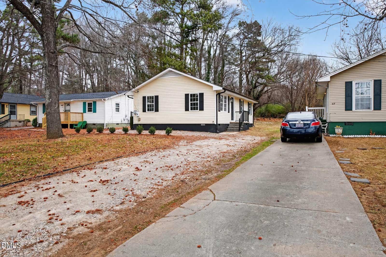 Single-story beige house with black shutters, a small porch, and gravel driveway. A blue car is parked on a concrete driveway to the right. Trees and neighboring houses are visible in the background.