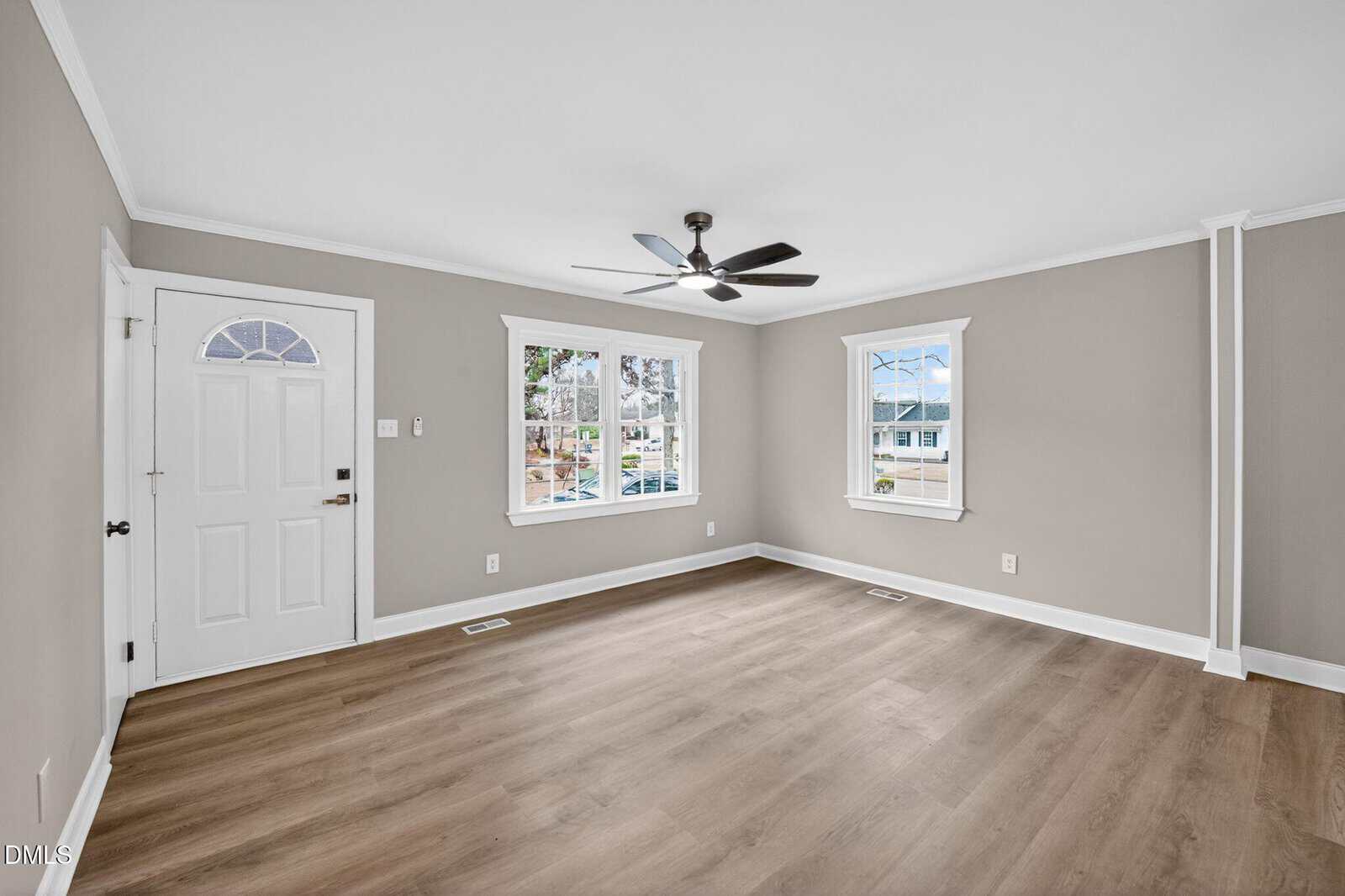 A bright, empty room with light wood flooring, gray walls, white trim, two windows, a white front door, and a black ceiling fan with lights. Outdoor houses and trees are visible through the windows.