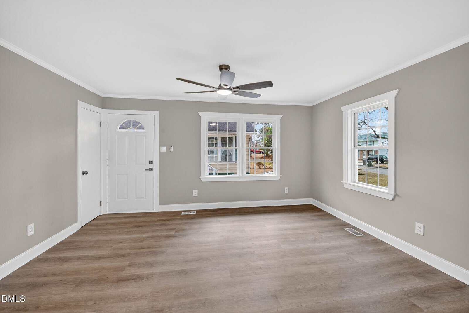 A bright, empty living room with light wood floors, gray walls, white trim, two windows, a white front door, and a ceiling fan with lights.