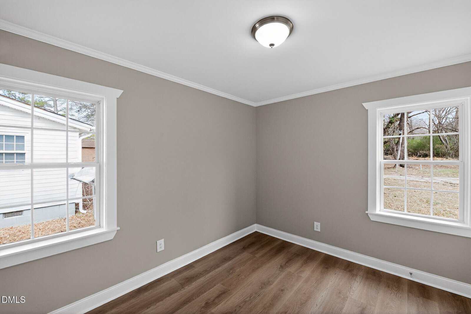 Empty room with beige walls, white trim, two windows showing an outside view, wood-style flooring, and a ceiling light fixture. Room appears clean and well-lit with natural daylight.