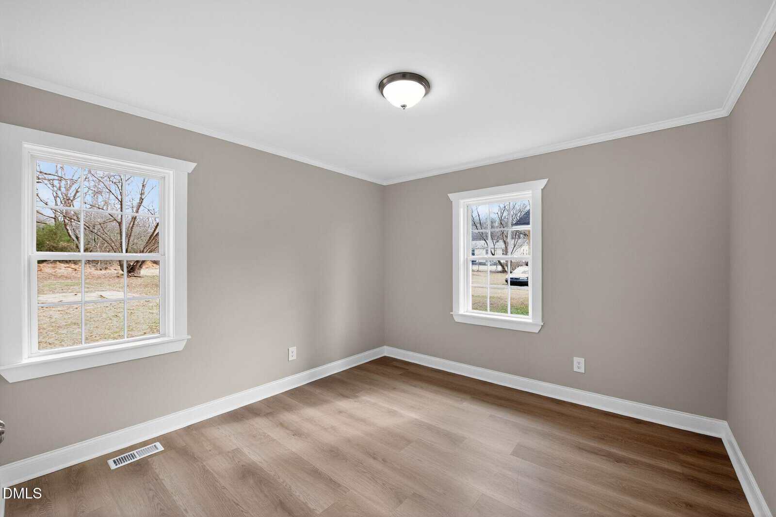 Empty room with light brown wood flooring, beige walls, white trim, and two windows letting in natural light. A ceiling light fixture is centered on the white ceiling. Outside, some grass and trees are visible.