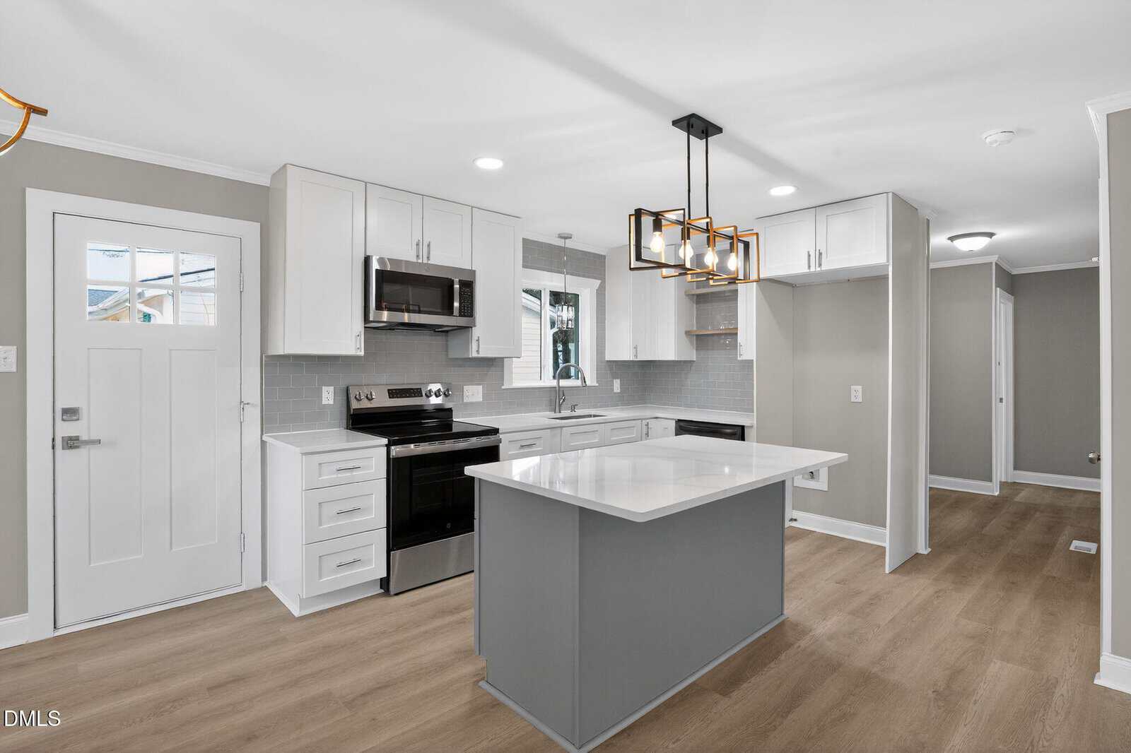 Modern kitchen with white cabinets, stainless steel appliances, a center island with white countertop, light gray walls, light wood flooring, and geometric pendant lighting over the island. Door and hallway are visible.