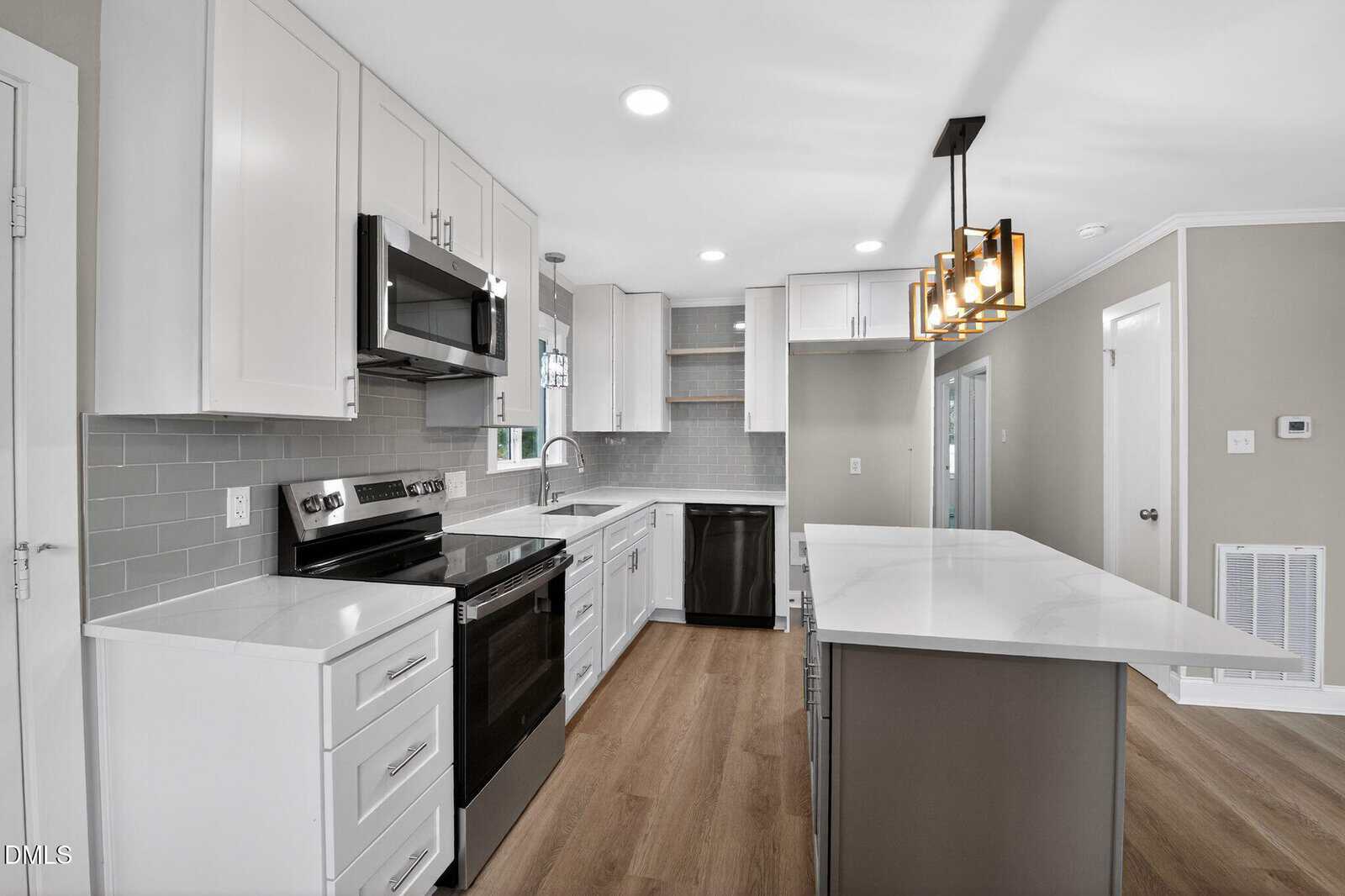 Modern kitchen with white cabinets, stainless steel appliances, gray subway tile backsplash, large island with white countertop, wood flooring, and a rectangular chandelier. Walls are light beige with white trim.