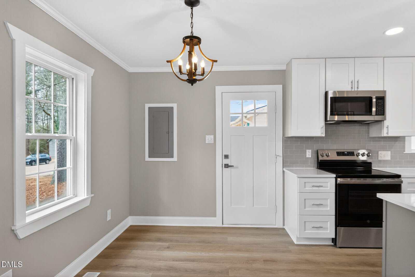 Modern kitchen with light wood floors, white cabinets, stainless steel appliances, gray backsplash, a window on the left, and a white door in the center. A decorative ceiling light hangs above.