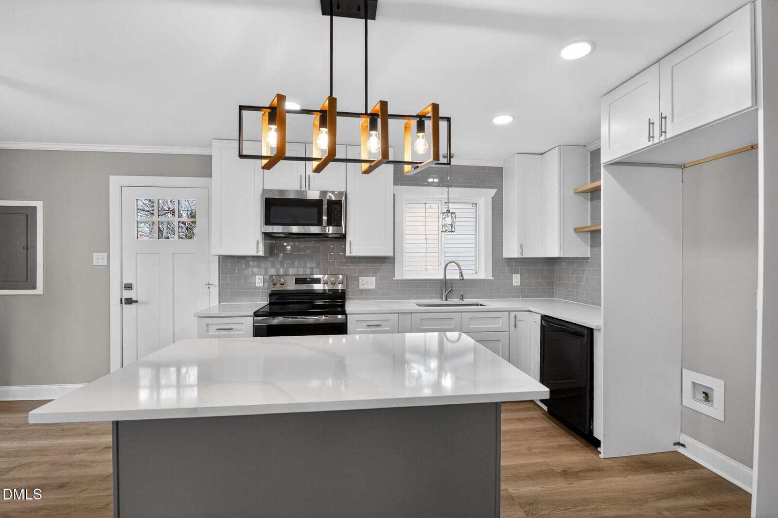 Modern kitchen with white cabinets, stainless steel appliances, a large island, pendant lighting, and wood flooring. There is space for a washer and dryer on the right and a window above the sink.