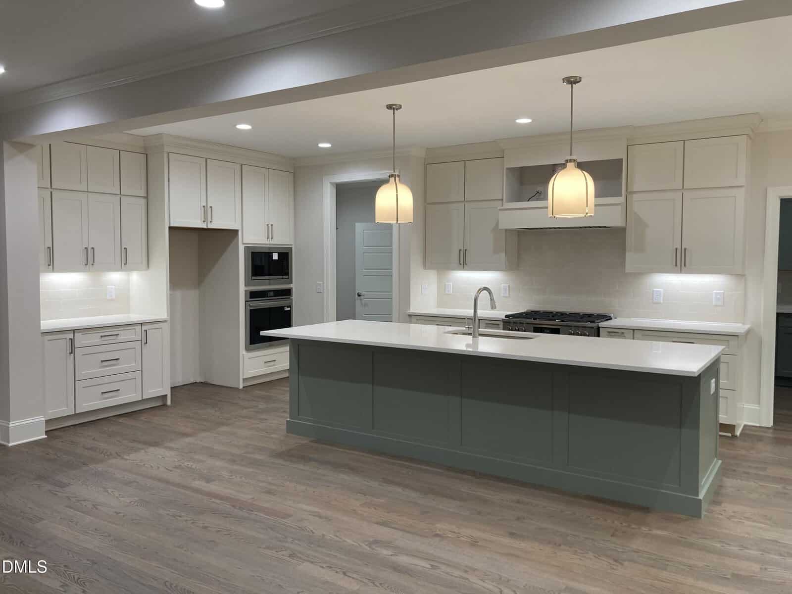 A modern kitchen with white cabinets, a large green island with a white countertop, double wall ovens, a built-in stove, pendant lights, and wood flooring. The space is well-lit with recessed and pendant lighting.