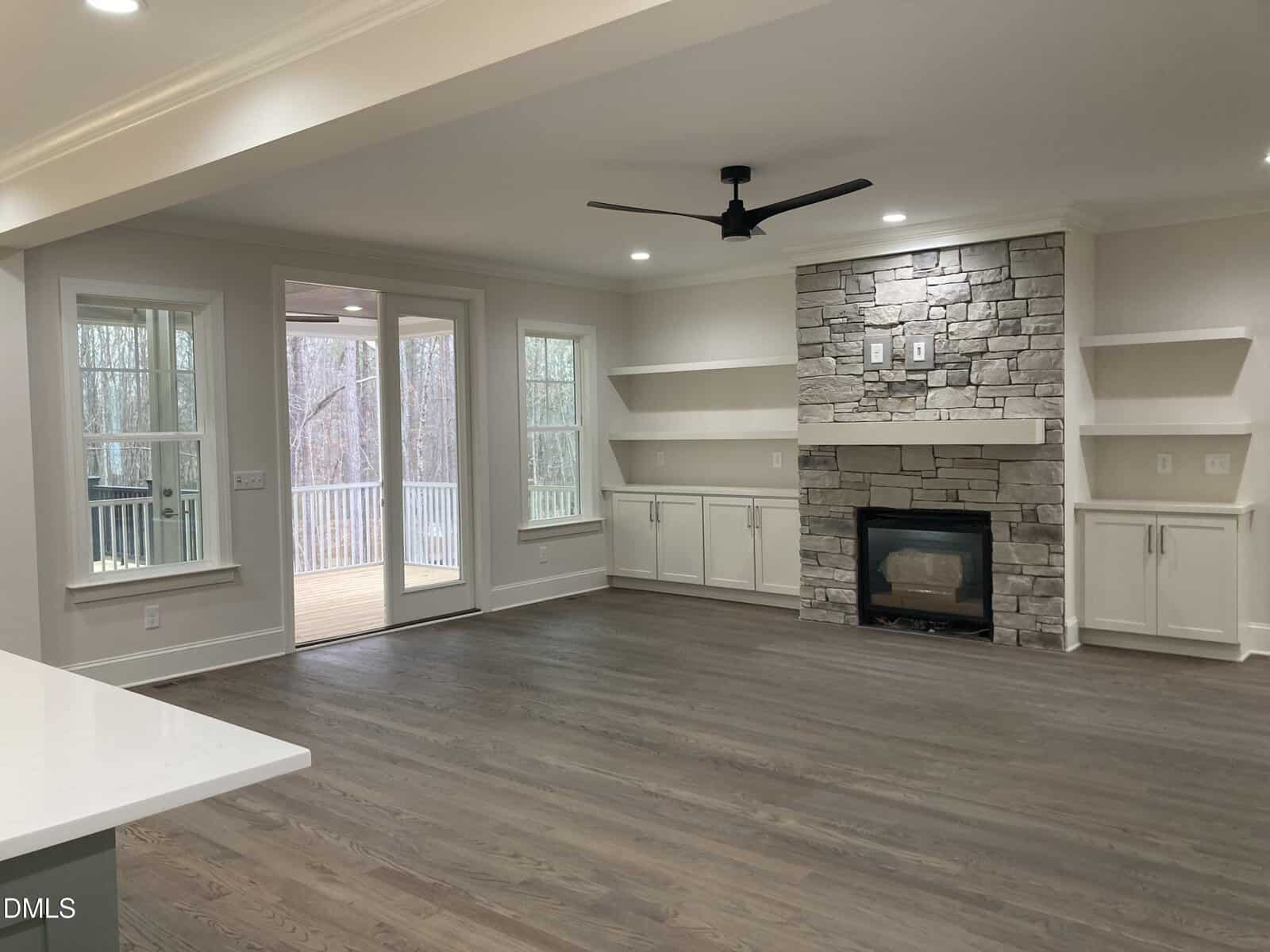 A spacious living room with light-colored walls and hardwood floors, featuring a stone fireplace with built-in shelves and cabinets on both sides, large windows, and sliding doors leading to a covered porch.