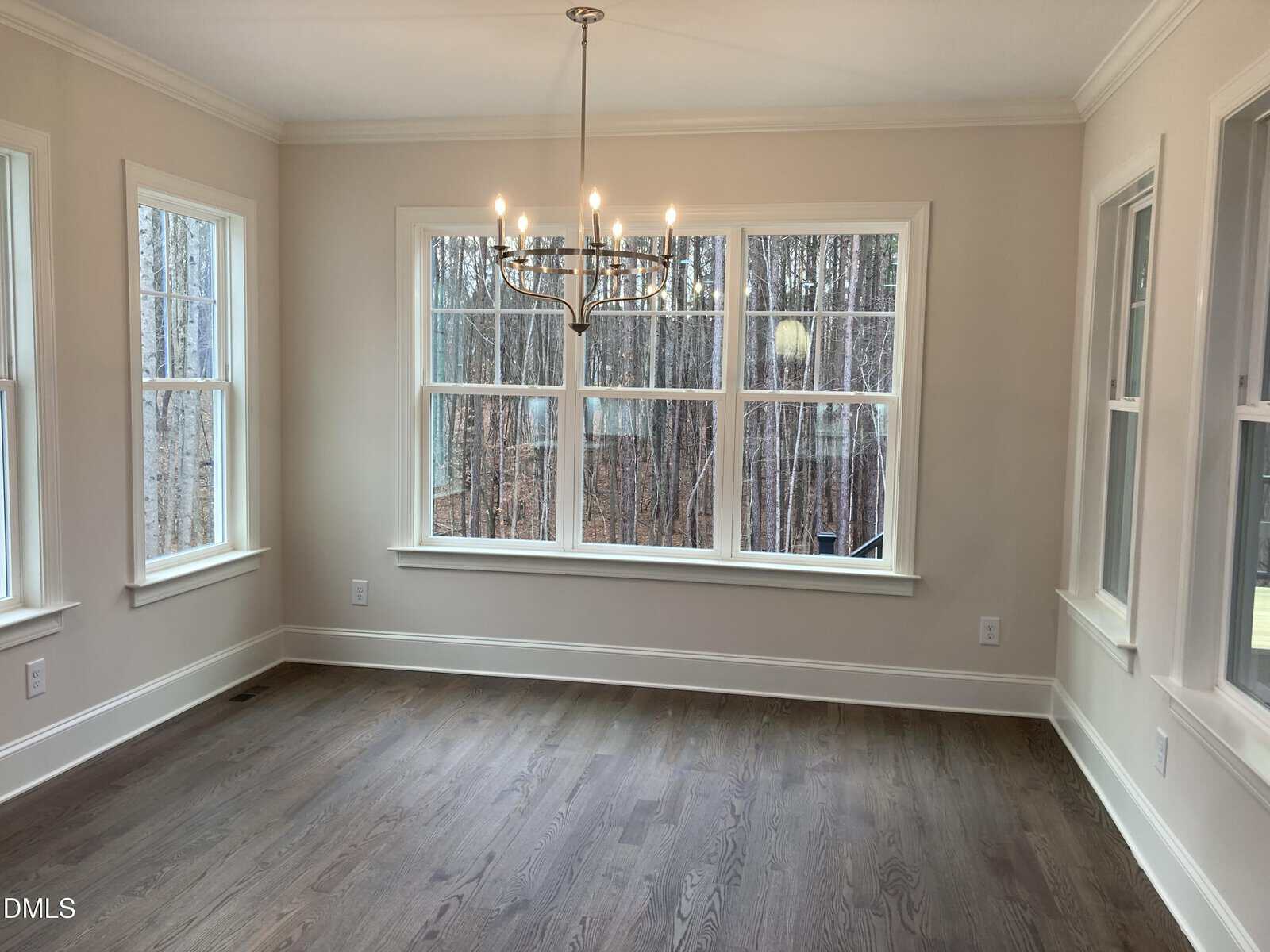 A bright, empty room with hardwood floors, beige walls, large windows on three sides, and a modern chandelier hanging from the ceiling. Leafless trees are visible through the windows.