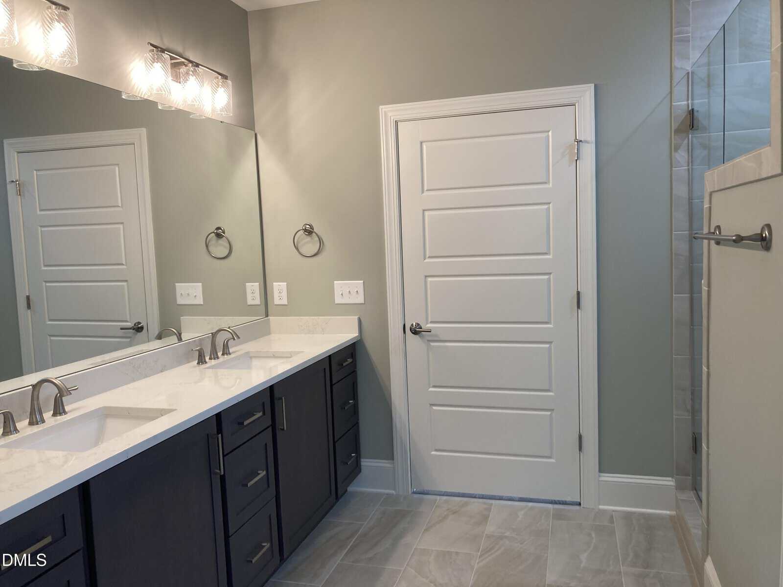 Modern bathroom with double sinks, dark cabinets, a large mirror, light fixtures above, a white door, tiled floor, and part of a glass shower visible on the right.