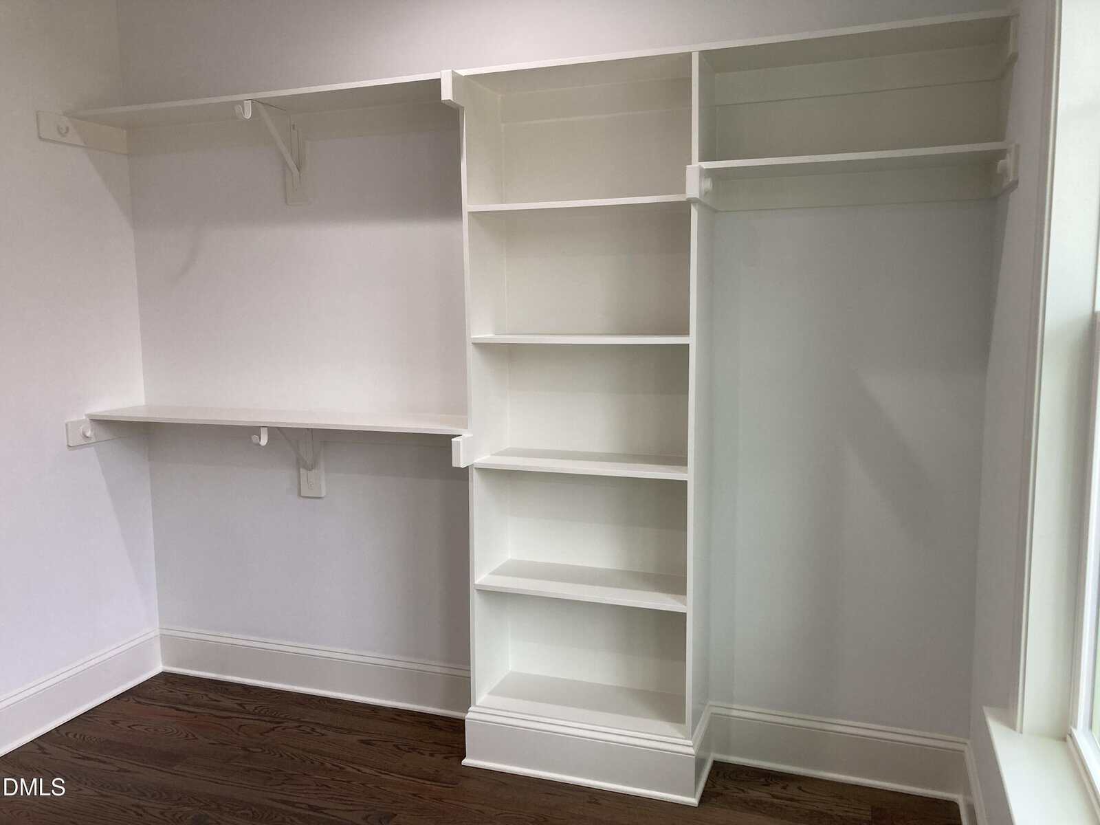 Walk-in closet with white built-in shelves, a hanging rod, and a small desk or counter area, with hardwood floors and white walls.