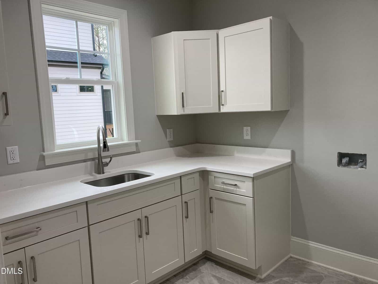 Modern kitchen with white cabinets, a stainless steel sink under a window, light gray walls, and no appliances installed. The countertop is clean and there are no visible decorations.