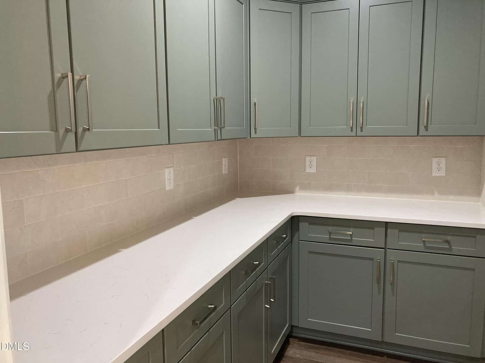 A kitchen corner with light green cabinets, white countertops, beige subway tile backsplash, multiple drawers, and several electrical outlets on the wall.