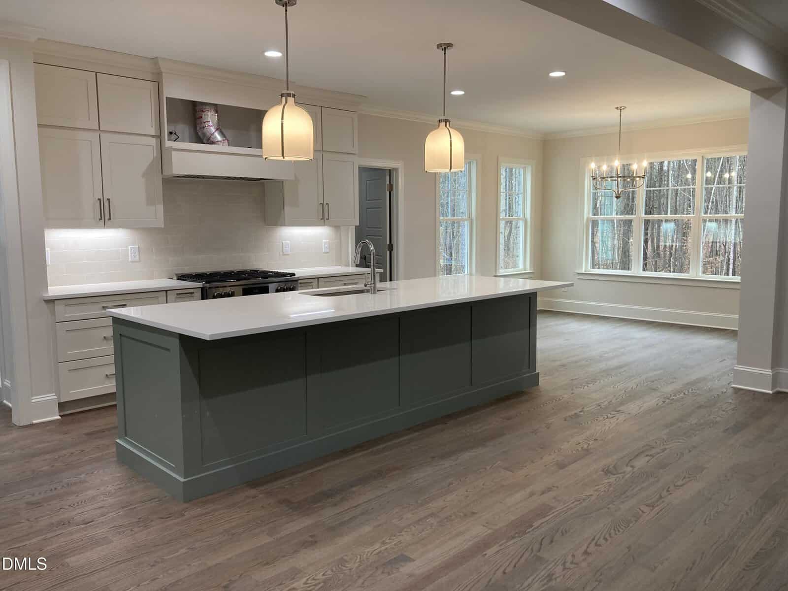 Modern kitchen with white cabinets, a green island with a sink, pendant lights, light wood floors, and an adjacent dining area with large windows and a chandelier.