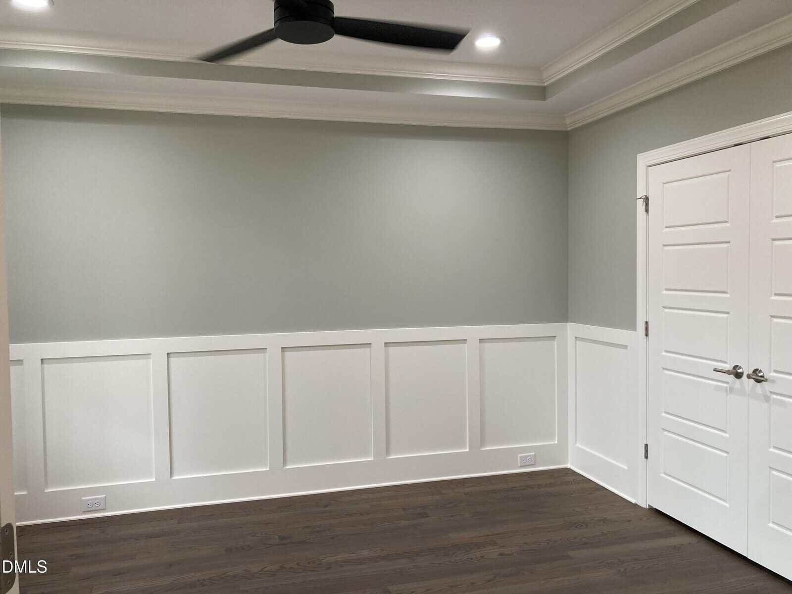 A room with light gray walls and white wainscoting, dark wood flooring, a white double door with silver handles, and a black ceiling fan with recessed lighting.