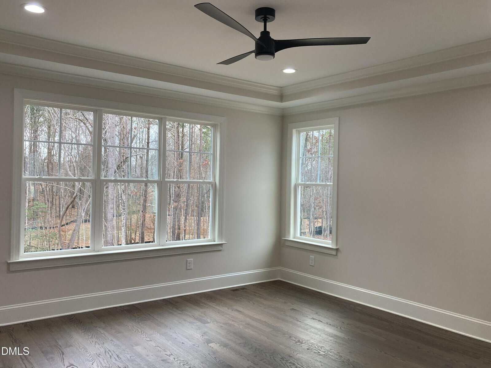 An empty room with light-colored walls, large windows showing trees outside, a dark ceiling fan, and wood flooring. Recessed lighting is on in the ceiling.