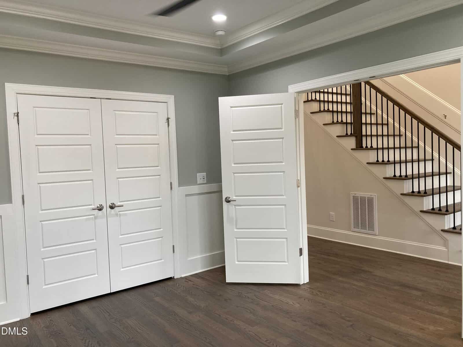A room with light gray walls, white trim, and dark hardwood floors; two closed double doors and one open single door are visible, leading to a hallway with a staircase featuring wooden steps and a black railing.