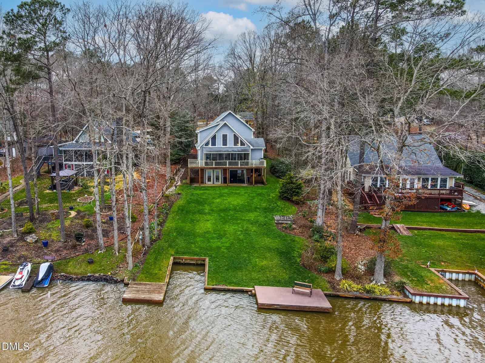 A two-story house with large windows and a deck overlooks a grassy lawn leading to a wooden dock on a lake. Neighboring houses and trees surround the property. The sky is partly cloudy.