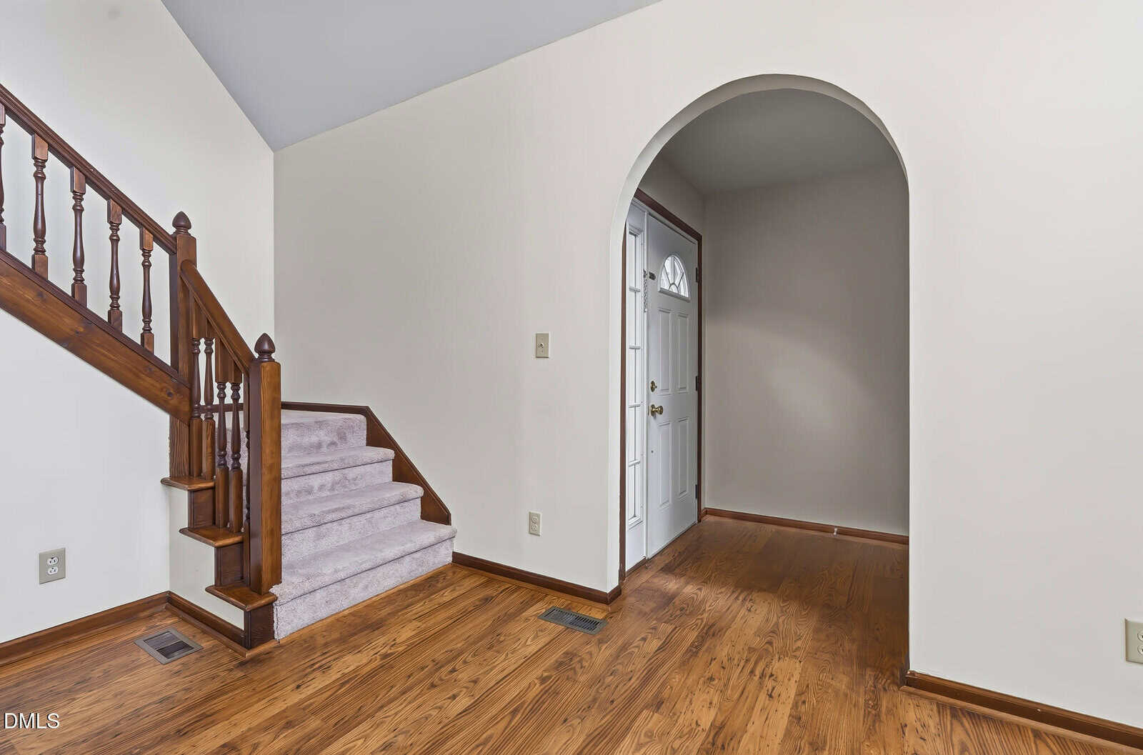 Staircase with wooden railing and carpeted steps next to an archway leading to a hallway and a white front door, with wood flooring throughout the space.