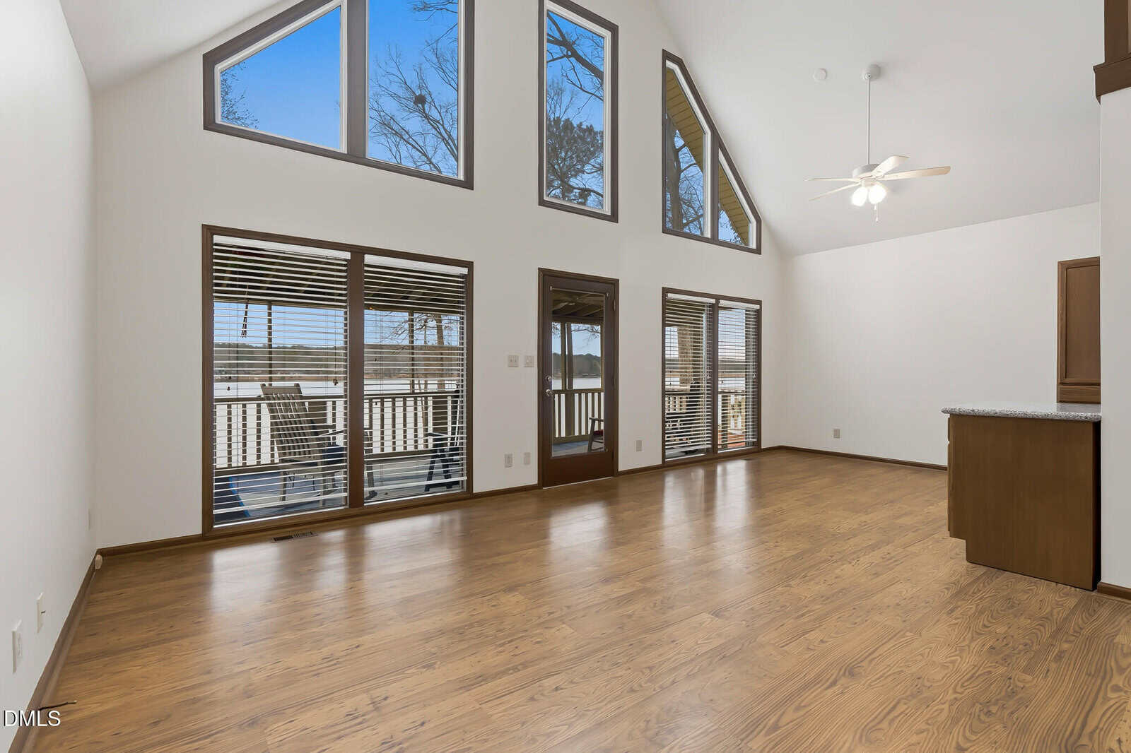 Spacious, empty living room with wood flooring, large windows offering outdoor views, a ceiling fan with lights, and an open kitchen area with granite countertops visible to the right.
