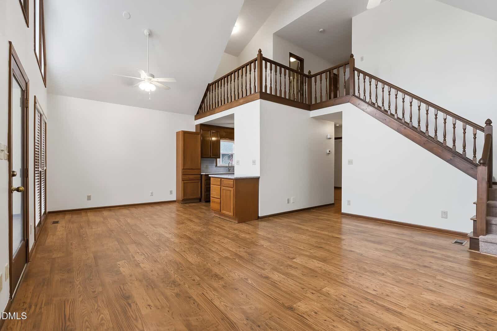 A spacious living area with wood flooring, a ceiling fan, and a vaulted ceiling. An open staircase with wooden railings leads to a loft area, and the kitchen is visible below with wooden cabinets.