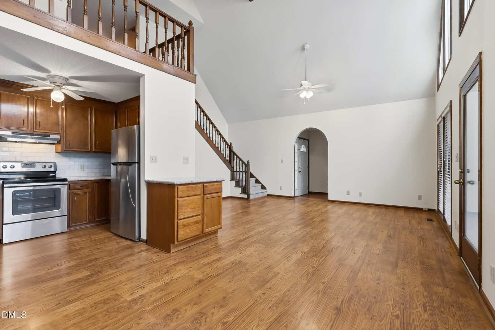 A spacious room with wood floors, an open kitchen featuring stainless steel appliances, wooden cabinets, a white countertop, and a loft area above. Stairs, ceiling fans, and large windows are visible.