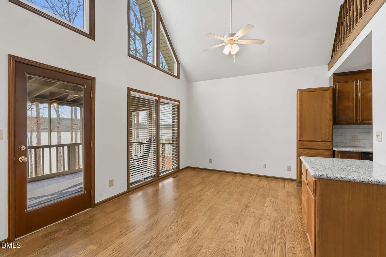 A bright, empty room with wood floors, large windows, a ceiling fan, a glass door to a balcony, and wood cabinets with granite countertops on the right. Loft railing visible above the kitchen area.