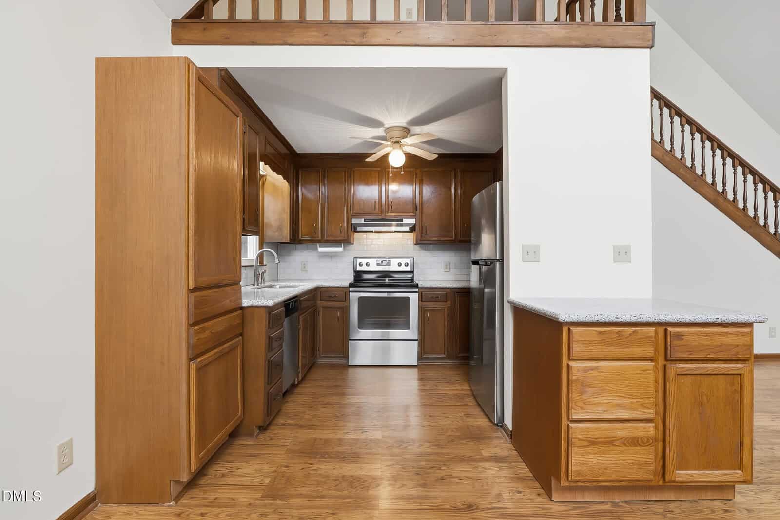 Kitchen with wooden cabinets, stainless steel appliances, a ceiling fan, granite countertops, and wood flooring. An open staircase and loft area are visible above and alongside the kitchen.