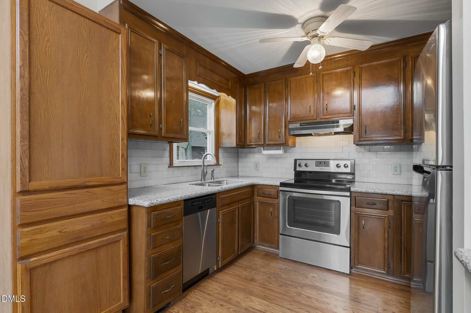 A kitchen with wooden cabinets, stainless steel appliances, a ceiling fan, white tile backsplash, and hardwood floors. There is a window above the sink and granite countertops throughout.